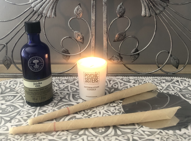 Two ear candles laid on a patterned tile beside a lit candle and a blue-glass bottle of jojoba oil
