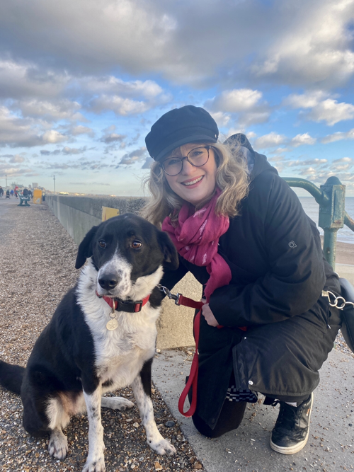 Paula outdoors on a seafront promenade, crouching with her arm around a black-and-white collie
