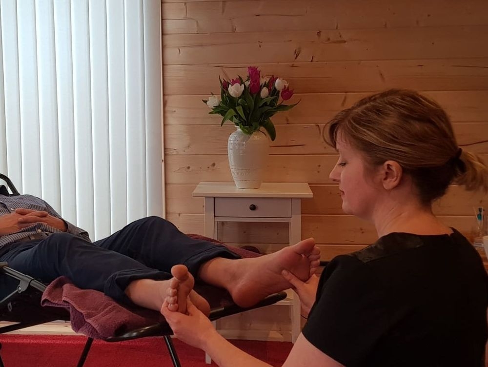 Paula seated at the end of a treatment couch, working with both hands on a client's feet in a pine-panelled room with tulips on the cabinet